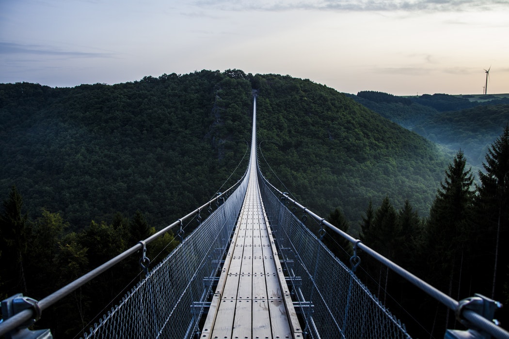 Dark green forest atop misty hills, with a long wood and steel bridge between them. Cross the bridge to Achieve your Dreams in Network Marketing