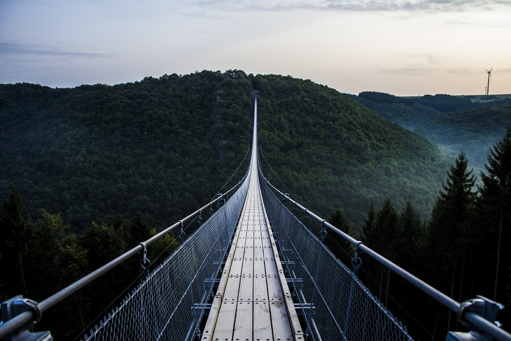 Dark green forest atop misty hills, with a long wood and steel bridge between them. Cross the bridge to Achieve your Dreams in Network Marketing